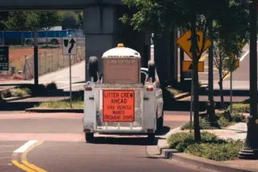 White utility vehicle with a sign "Litter Crew Ahead, This Vehicle Makes Frequent Stops" parked on a street under a bridge near trees and road signs.