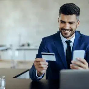 A man in a blue suit sits smiling at a desk, holding a credit card and smartphone, suggesting online shopping. A blurred office background is visible.