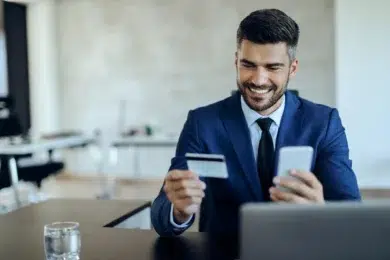 A man in a blue suit sits smiling at a desk, holding a credit card and smartphone, suggesting online shopping. A blurred office background is visible.