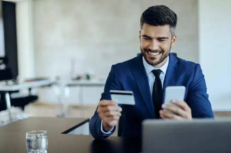 A man in a blue suit sits smiling at a desk, holding a credit card and smartphone, suggesting online shopping. A blurred office background is visible.