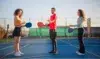 Three people holding paddles and balls on a pickleball court, facing each other. The scene is sunny and lighthearted with a clear sky.