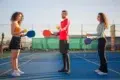 Three people holding paddles and balls on a pickleball court, facing each other. The scene is sunny and lighthearted with a clear sky.