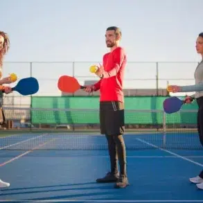 Three people holding paddles and balls on a pickleball court, facing each other. The scene is sunny and lighthearted with a clear sky.