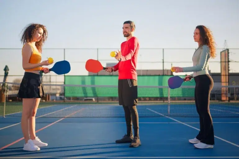 Three people holding paddles and balls on a pickleball court, facing each other. The scene is sunny and lighthearted with a clear sky.