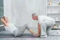 An older woman doing sit-ups on a yoga mat indoors, assisted by an older man kneeling and holding her feet. They are both smiling and wearing casual workout clothes, creating a supportive and positive atmosphere.