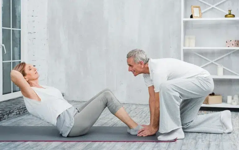 An older woman doing sit-ups on a yoga mat indoors, assisted by an older man kneeling and holding her feet. They are both smiling and wearing casual workout clothes, creating a supportive and positive atmosphere.