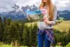 A woman stands amidst a lush, mountainous landscape holding a colorful map. She wears a gray sweatshirt and jeans, conveying a sense of adventure.