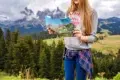 A woman stands amidst a lush, mountainous landscape holding a colorful map. She wears a gray sweatshirt and jeans, conveying a sense of adventure.
