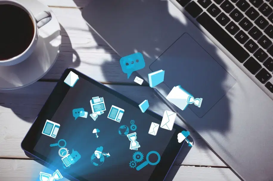 Tablet and laptop on a desk with digital icons of emails, documents, and communication floating above, next to a cup of coffee.