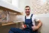 A smiling plumber in blue overalls gives a thumbs-up while holding a wrench under a bathroom sink. The setting is clean and modern, conveying satisfaction.