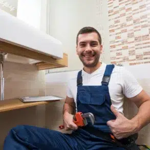 A smiling plumber in blue overalls gives a thumbs-up while holding a wrench under a bathroom sink. The setting is clean and modern, conveying satisfaction.