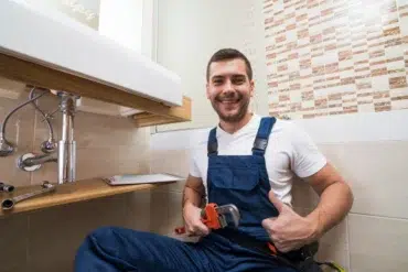 A smiling plumber in blue overalls gives a thumbs-up while holding a wrench under a bathroom sink. The setting is clean and modern, conveying satisfaction.