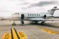 Private jet on a tarmac under a cloudy sky. The jet's door is open, with a small staircase leading up. Yellow tarmac markings are visible.