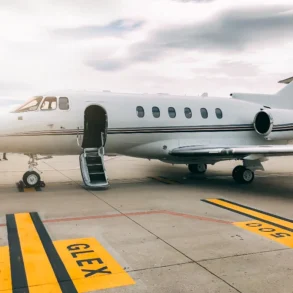Private jet on a tarmac under a cloudy sky. The jet's door is open, with a small staircase leading up. Yellow tarmac markings are visible.