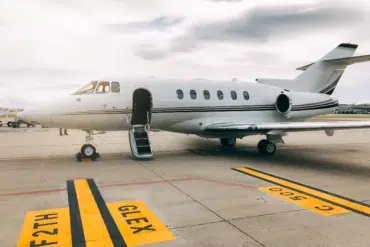 Private jet on a tarmac under a cloudy sky. The jet's door is open, with a small staircase leading up. Yellow tarmac markings are visible.