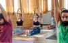 A group of people in a bright room practicing seated yoga. They sit cross-legged with arms raised. The atmosphere is calm and focused, with natural light.