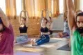A group of people in a bright room practicing seated yoga. They sit cross-legged with arms raised. The atmosphere is calm and focused, with natural light.