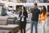 A couple holding hands stands with a clipboard-holding saleswoman in a modern furniture store, surrounded by sofas. The mood is professional yet relaxed.
