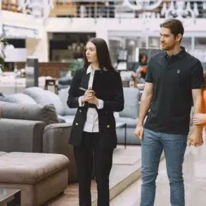 A couple holding hands stands with a clipboard-holding saleswoman in a modern furniture store, surrounded by sofas. The mood is professional yet relaxed.