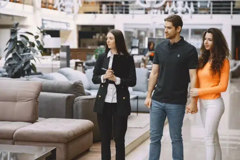 A couple holding hands stands with a clipboard-holding saleswoman in a modern furniture store, surrounded by sofas. The mood is professional yet relaxed.
