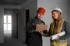 Two construction workers in safety gear review blueprints on a clipboard in an unfinished building. The mood is focused and professional.