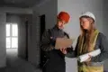Two construction workers in safety gear review blueprints on a clipboard in an unfinished building. The mood is focused and professional.