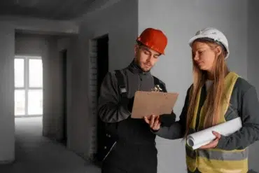 Two construction workers in safety gear review blueprints on a clipboard in an unfinished building. The mood is focused and professional.