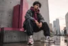 Young man in urban streetwear sits on a concrete ledge, exuding confidence. Skyscrapers and wet pavement suggest a moody cityscape backdrop.