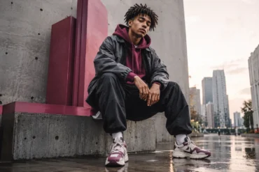 Young man in urban streetwear sits on a concrete ledge, exuding confidence. Skyscrapers and wet pavement suggest a moody cityscape backdrop.