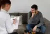 Therapist taking notes while a man in a gray sweater sits on a couch, appearing thoughtful during a counseling session.