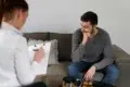 Therapist taking notes while a man in a gray sweater sits on a couch, appearing thoughtful during a counseling session.