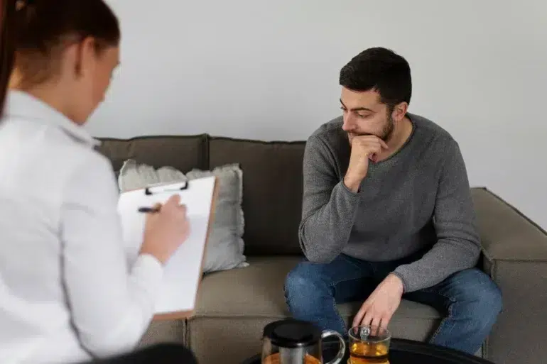 Therapist taking notes while a man in a gray sweater sits on a couch, appearing thoughtful during a counseling session.