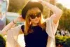 A woman in a black dress and beige shawl adjusts a wide-brimmed hat, wearing sunglasses. She stands outdoors, bathed in warm sunlight, exuding a relaxed vibe.