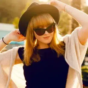 A woman in a black dress and beige shawl adjusts a wide-brimmed hat, wearing sunglasses. She stands outdoors, bathed in warm sunlight, exuding a relaxed vibe.