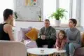 Therapist listens to a family with a child drawing on a small table in a bright living room with plants and abstract art.