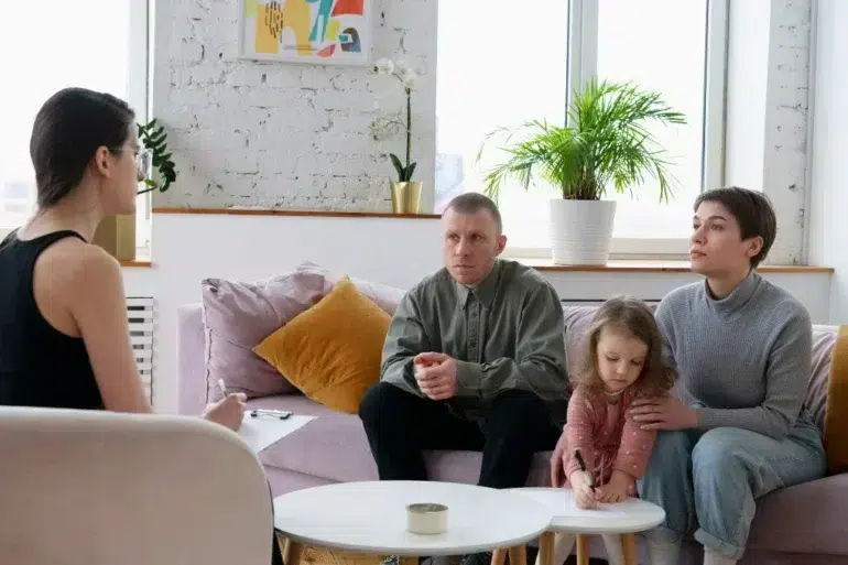 Therapist listens to a family with a child drawing on a small table in a bright living room with plants and abstract art.