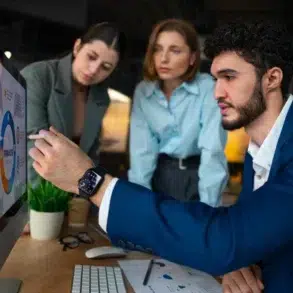 A man in a blue suit points to data on a computer screen, discussing with two women who focus attentively. The atmosphere is collaborative.
