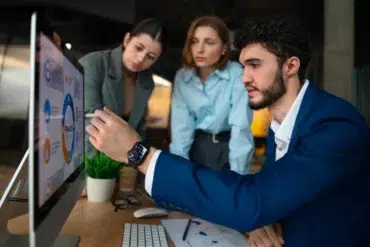 A man in a blue suit points to data on a computer screen, discussing with two women who focus attentively. The atmosphere is collaborative.