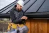 A construction worker in a white hard hat and gray jacket examines a piece of material on a roof. He wears a tool belt, earphones, and stands near wood siding.