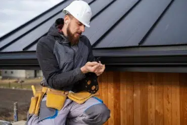A construction worker in a white hard hat and gray jacket examines a piece of material on a roof. He wears a tool belt, earphones, and stands near wood siding.