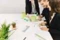 Three colleagues in a meeting, smiling and discussing designs at a white table with notebooks, green pencils, and potted plants for a creative atmosphere.