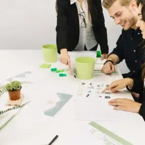 Three colleagues in a meeting, smiling and discussing designs at a white table with notebooks, green pencils, and potted plants for a creative atmosphere.