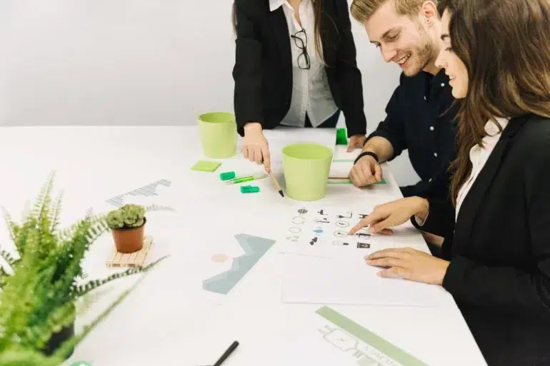 Three colleagues in a meeting, smiling and discussing designs at a white table with notebooks, green pencils, and potted plants for a creative atmosphere.