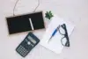 A flat lay on a light wooden surface features a small blackboard with chalk, a scientific calculator, a spiral notebook with a pen, eyeglasses, and a small succulent plant, conveying a study or planning theme.
