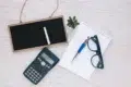 A flat lay on a light wooden surface features a small blackboard with chalk, a scientific calculator, a spiral notebook with a pen, eyeglasses, and a small succulent plant, conveying a study or planning theme.