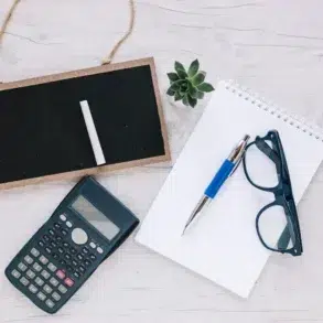 A flat lay on a light wooden surface features a small blackboard with chalk, a scientific calculator, a spiral notebook with a pen, eyeglasses, and a small succulent plant, conveying a study or planning theme.