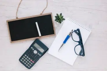 A flat lay on a light wooden surface features a small blackboard with chalk, a scientific calculator, a spiral notebook with a pen, eyeglasses, and a small succulent plant, conveying a study or planning theme.