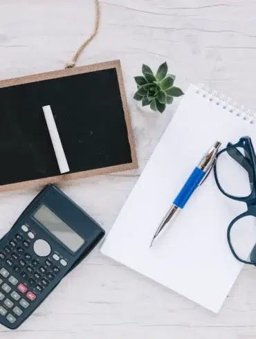 A flat lay on a light wooden surface features a small blackboard with chalk, a scientific calculator, a spiral notebook with a pen, eyeglasses, and a small succulent plant, conveying a study or planning theme.