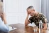 Man in camouflage shirt holding his head, appearing distressed, during a conversation with a woman at a wooden table with a notebook and water.