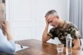 Man in camouflage shirt holding his head, appearing distressed, during a conversation with a woman at a wooden table with a notebook and water.